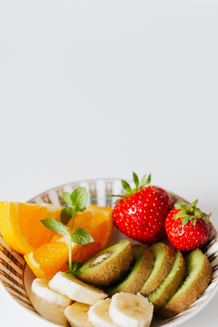 our-story Close-up of a fresh fruit bowl with kiwi, strawberries, bananas, and oranges on a white background.