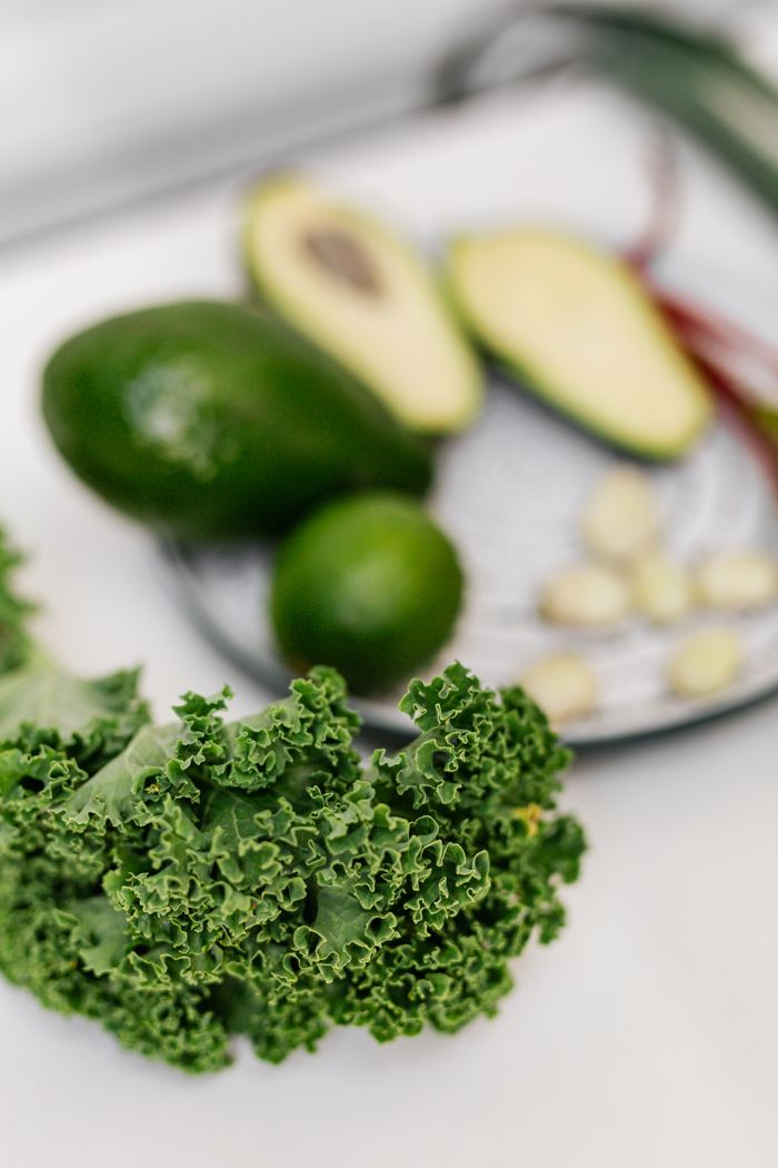 A close-up of fresh kale with avocado, lime, and garlic on a white surface.